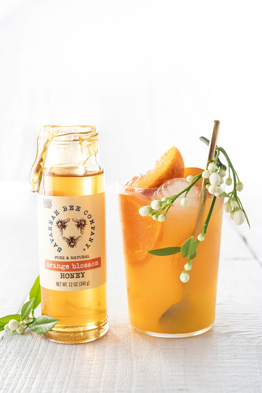 A fuzzy navel drink with fruits and garnish next to a 12 ounce jar of orange blossom honey.